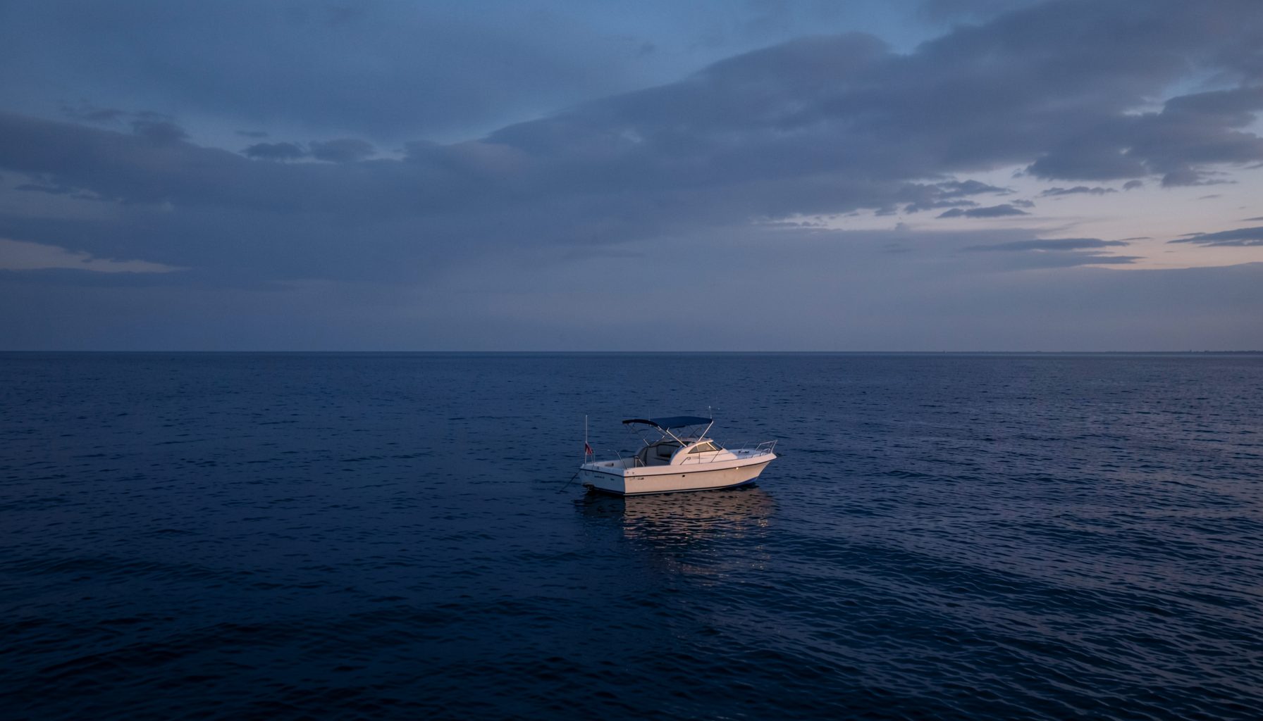 Ruhiges dunkelblaues Meer mit einem kleinen weißen Sportboot bei sanftem Abendlicht, gedämpfte Wolken, ruhige und vertrauensvolle Stimmung